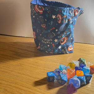 May include: A blue fabric dice bag with a Captain America pattern. The bag is filled with various colored dice, including blue, red, green, orange, and white.
