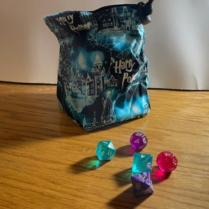 May include: A blue and black fabric dice bag with a Harry Potter design. The bag is sitting on a wooden table with five dice scattered around it. The dice are various colors, including green, purple, and red.