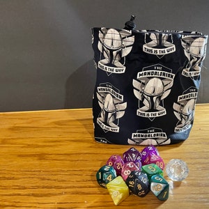 May include: A black fabric dice bag with a repeating pattern of The Mandalorian's helmet and the phrase "This is the way". The bag is filled with a set of colorful dice and a clear d20.