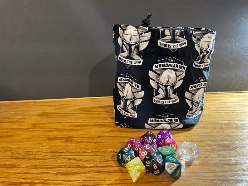 May include: A black fabric dice bag with a repeating pattern of The Mandalorian's helmet and the phrase "This is the way". The bag is filled with a set of colorful dice and a clear d20.