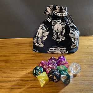 May include: A black fabric dice bag with a white print of "The Mandalorian" and "This is the way". The bag is filled with colorful dice and a clear d20.