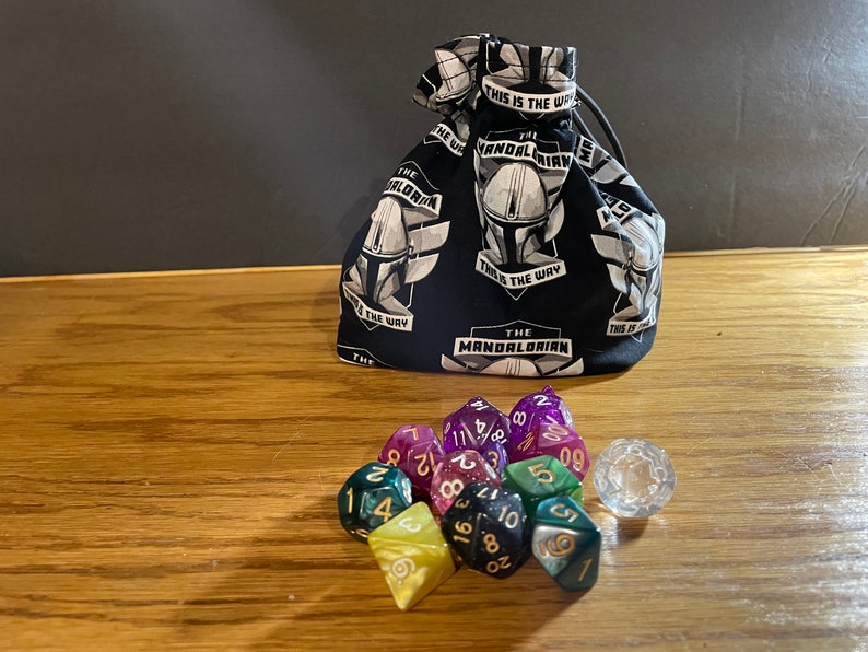 May include: A black fabric dice bag with a white print of "The Mandalorian" and "This is the way". The bag is filled with colorful dice and a clear d20.