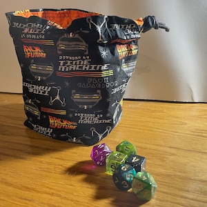 May include: A black drawstring bag with a repeating pattern of the DeLorean time machine from the Back to the Future movies. The bag is filled with green and purple dice.