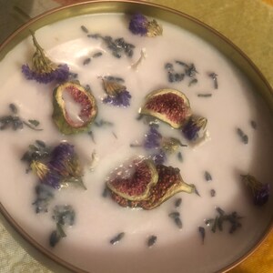 Puede incluir: Una vela blanca con lavanda seca y rodajas de higo incrustadas en la cera. La vela está en un recipiente dorado.