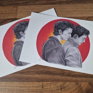 May include: Two rectangular art prints featuring a graphite-style illustration of two individuals standing side-by-side in profile. The figures are set against a vibrant circular background that transitions from a deep red outer edge to a bright yellow centre.
