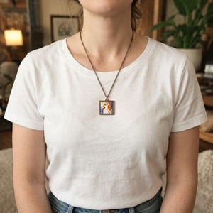 May include: A square pendant necklace with a bronze-colored chain. The pendant features a colorful image within a bronze-colored square frame. The person is wearing a white t-shirt and blue jeans.