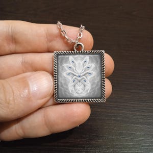 May include: Square pendant necklace with a silver chain. The pendant features a detailed illustration of a white, winged creature with multiple blue eyes against a gray background. The pendant is framed with a decorative silver border.