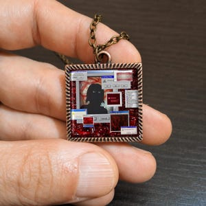 May include: A square pendant necklace with a copper-colored frame and a dark chain. The pendant features a digital art design with a silhouetted figure, computer windows, and a red background. The pendant is held in a hand.