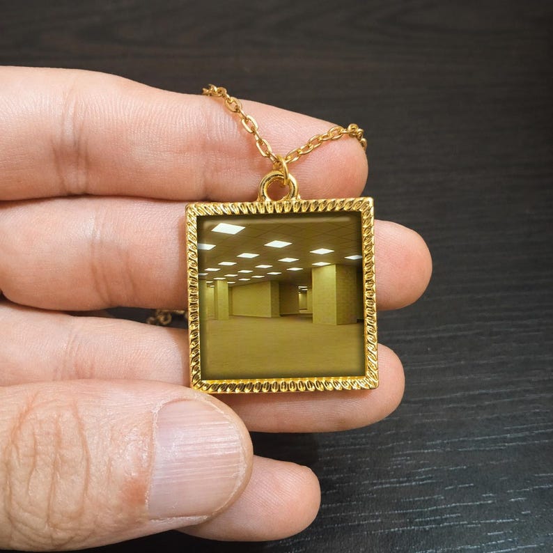 May include: Gold-toned square pendant necklace featuring a surreal, yellow-toned image of a liminal space. The pendant has a textured border and hangs from a delicate chain. The necklace is held in a hand against a dark background.
