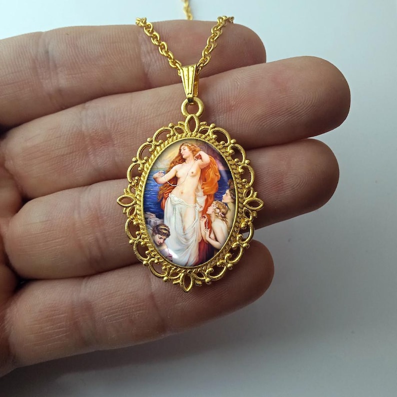 May include: Gold-toned pendant necklace featuring an oval-shaped miniature painting of a classical scene. The painting depicts a nude woman with red hair, surrounded by figures, set against a blue sea backdrop. The pendant has a decorative filigree frame.