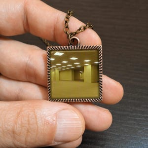 May include: A bronze-toned square pendant necklace featuring a surreal image of a yellow-lit, empty room. The pendant has a textured border and hangs from a matching chain. The necklace is held up against a dark background.