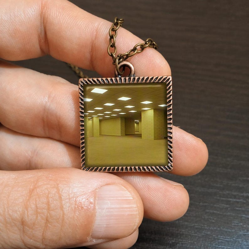 May include: A bronze-toned square pendant necklace featuring a surreal image of a yellow-lit, empty room. The pendant has a textured border and hangs from a matching chain. The necklace is held up against a dark background.