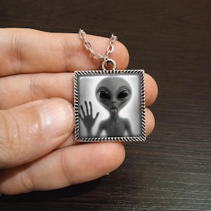 May include: A silver-toned necklace featuring a square pendant with a gray alien graphic. The alien has large black eyes and is sticking out its tongue. The pendant is framed with a decorative border and hangs from a delicate chain.