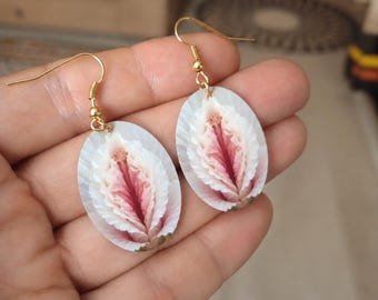 May include: A pair of gold-tone dangle earrings. Each earring features an oval-shaped pendant with a detailed, layered design in shades of white and pink. The earrings are held by a hand.