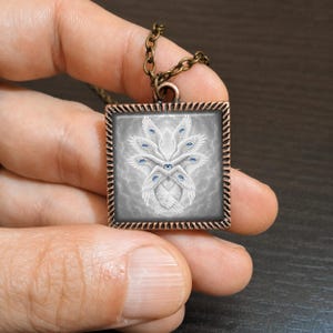 May include: Square pendant necklace with a bronze chain. The pendant features a detailed illustration of a white, winged creature with multiple blue eyes against a gray background. The pendant has a decorative border.