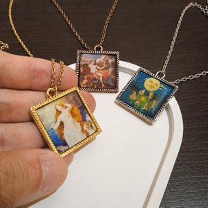 May include: Three square pendant necklaces with different art prints. One pendant has a gold frame and depicts a woman with long hair. Another has a copper frame with a scene of figures. The third has a silver frame with a moon and figures.