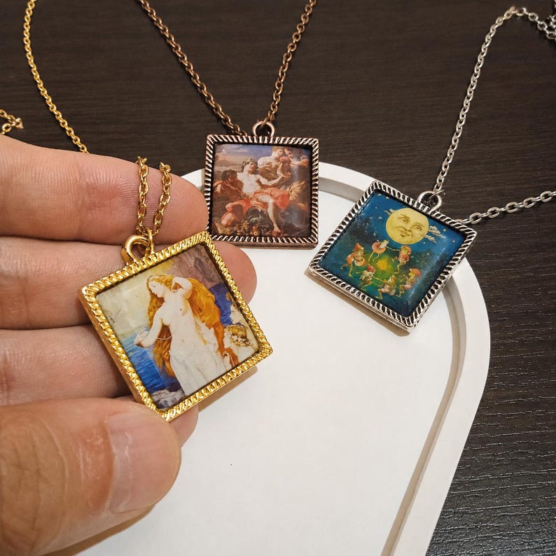 May include: Three square pendant necklaces with different art prints. One pendant has a gold frame and depicts a woman with long hair. Another has a copper frame with a scene of figures. The third has a silver frame with a moon and figures.