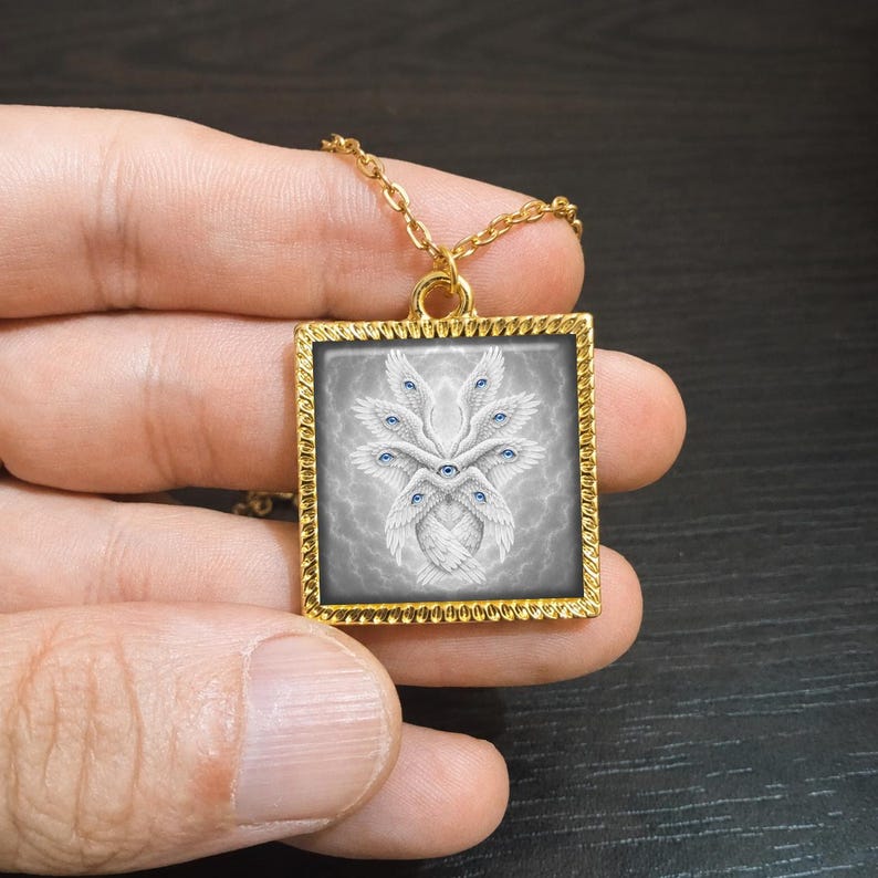 May include: Gold-toned square pendant necklace featuring a detailed illustration of a multi-eyed, winged creature with blue eyes and white feathers. The pendant is suspended from a delicate gold chain, creating a striking accessory.