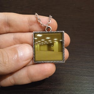 May include: Silver-toned square pendant necklace featuring a surreal, yellow-toned room scene. The pendant has a decorative border and hangs from a delicate chain. The image inside the pendant depicts a long hallway with evenly spaced lights.