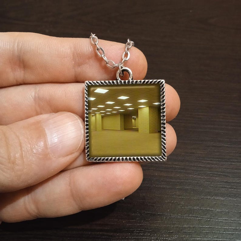May include: Silver-toned square pendant necklace featuring a surreal, yellow-toned room scene. The pendant has a decorative border and hangs from a delicate chain. The image inside the pendant depicts a long hallway with evenly spaced lights.