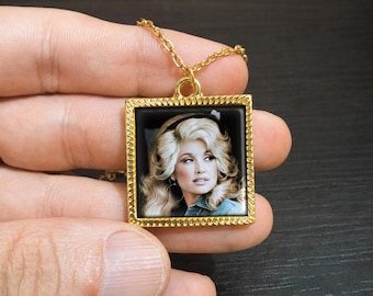 May include: A gold-toned square pendant necklace featuring a photographic portrait of a blonde person with voluminous hair. The pendant has a textured, rope-style border and is suspended from a matching gold-toned metal chain link necklace.