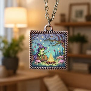 Può includere: Una collana con pendente quadrato con un'illustrazione stravagante di una rana con un cappello da mago che arrostisce un marshmallow su un falò. Il pendente ha una cornice color bronzo e pende da una catena abbinata. Lo sfondo presenta una scena della foresta.