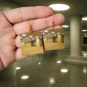 May include: A pair of square-shaped dangle earrings featuring a printed image of a yellow-toned, empty office hallway with fluorescent ceiling lights. The earrings are attached to gold-toned metal hooks and display a repeating architectural pattern.