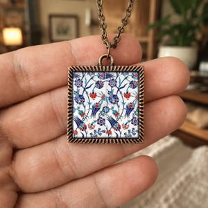 May include: A square pendant necklace with a bronze-colored frame. The pendant features a white background with a blue, red, and green floral pattern. The necklace chain is also bronze-colored.