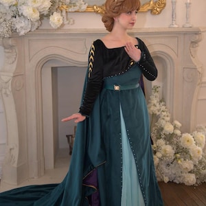 Op de afbeelding: Een vloerlange blauwgroene en zwarte japon met een vierkante halslijn, lange mouwen met gouden chevron-borduursels, een blauwgroene riem met een centrale gesp, een geplooide lichtblauwe onderrok, een lange blauwgroene cape en een gouden kroon.