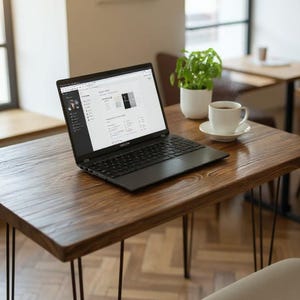 May include: A black laptop is open on a wooden table with black hairpin legs. A white teacup and saucer, and a small potted plant are also on the table. A white chair is in the foreground, and a cafe setting is in the background.