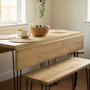 May include: A light-toned wooden dining table with a drop-leaf side and a matching wooden bench, both featuring thin black metal hairpin legs. The table surface holds a wooden bowl with a banana, a ceramic mug, and two woven placemats.
