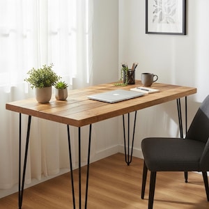 May include: A rectangular wooden desk with a natural grain finish and four thin black metal hairpin legs. On the desk surface sits a silver laptop, a small potted green plant, a smaller succulent, a black pencil holder, and a gray ceramic mug.