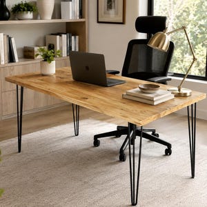 May include: A rectangular wooden desk with a natural grain finish and four black metal hairpin legs. The desk surface holds a dark gray laptop, a small potted green plant in a white ribbed ceramic pot, a stack of two books, and a gold desk lamp.