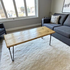 May include: A rectangular wooden coffee table with black hairpin legs. The table has a natural wood finish and sits on a white, shaggy rug. A gray sofa and a window are in the background.