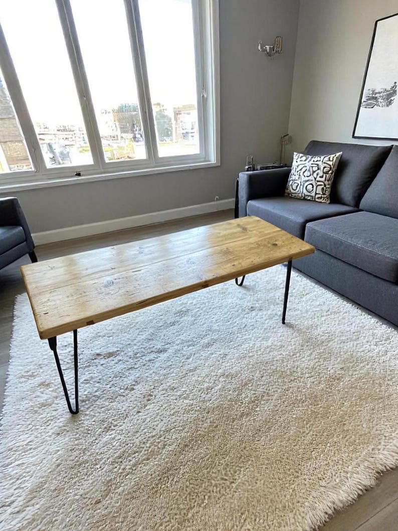 May include: A rectangular wooden coffee table with black hairpin legs. The table has a natural wood finish and sits on a white, shaggy rug. A gray sofa and a window are in the background.