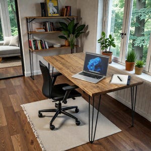 May include: A wooden desk with a natural grain finish and black metal hairpin legs, featuring a silver laptop, a spiral-bound notebook with a black pen, a black office chair, a tall bookshelf, and several potted green plants in a room.