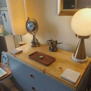 May include: A wooden dresser with a light blue painted frame and a natural wood top. Decorative items include a globe clock, a cat statue wearing glasses, a wooden tray, and a table lamp with a white globe shade.