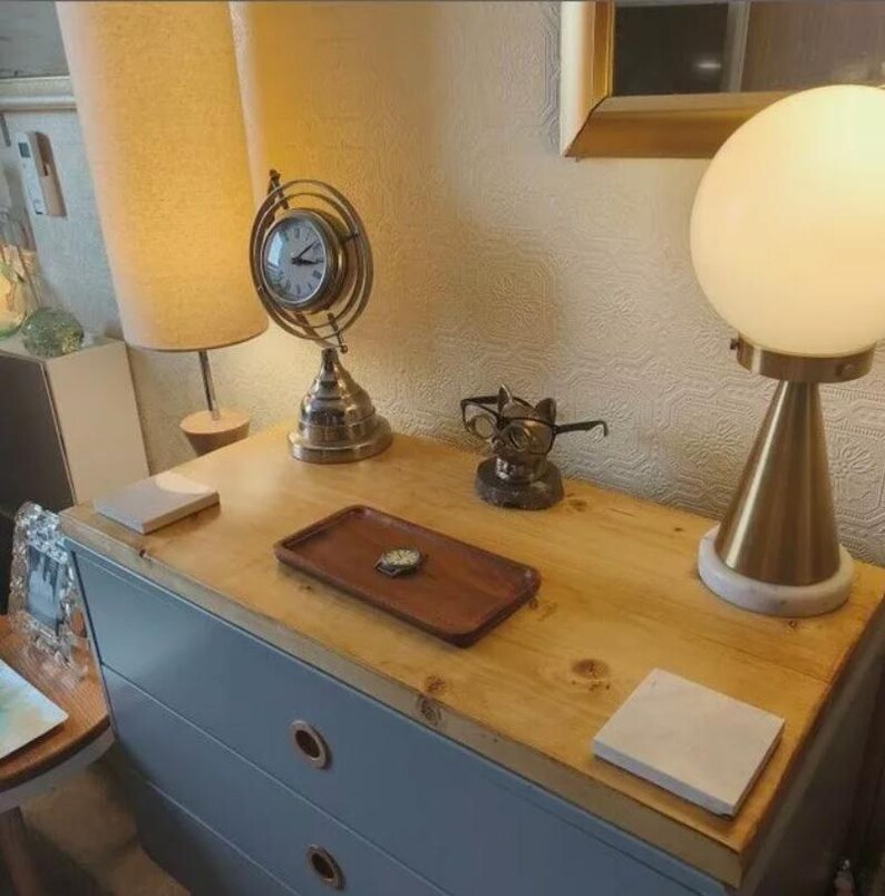 May include: A wooden dresser with a light blue painted frame and a natural wood top. Decorative items include a globe clock, a cat statue wearing glasses, a wooden tray, and a table lamp with a white globe shade.