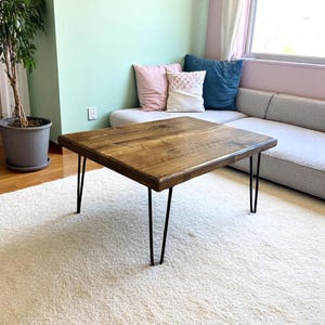 May include: Square wooden coffee table with a natural wood finish. The table has black hairpin legs. The table is approximately 30 inches wide and 16 inches tall. The table is placed on a white shag rug.