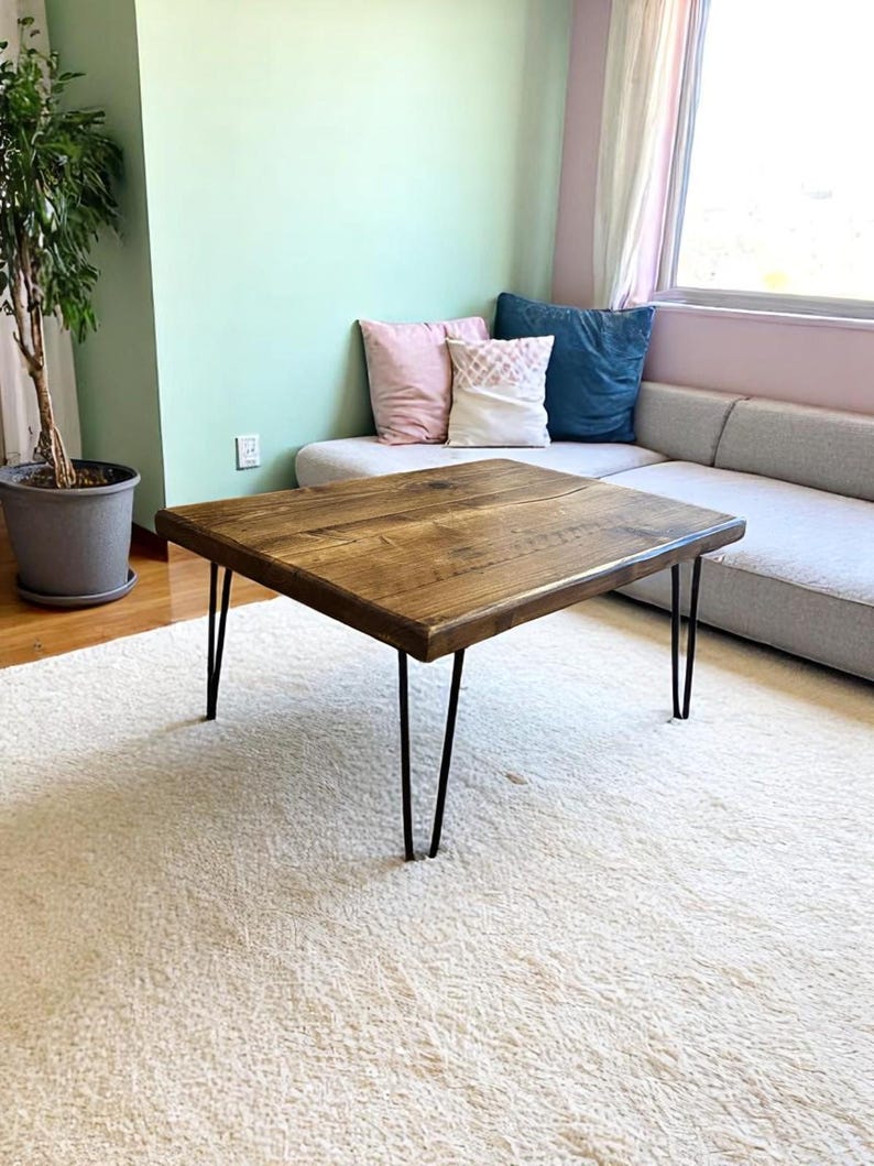 May include: Square wooden coffee table with a natural wood finish. The table has black hairpin legs. The table is approximately 30 inches wide and 16 inches tall. The table is placed on a white shag rug.