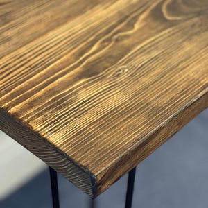 May include: A rectangular table featuring a thick, dark brown stained wooden top with a prominent natural grain pattern and texture. The tabletop is supported by thin, black metal hairpin legs that extend downward from the corners.
