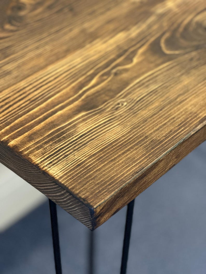 May include: A rectangular table featuring a thick, dark brown stained wooden top with a prominent natural grain pattern and texture. The tabletop is supported by thin, black metal hairpin legs that extend downward from the corners.