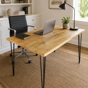 May include: A rectangular desk with a natural light brown wood grain top and four thin black metal hairpin legs. The desk surface holds a silver laptop, a black notebook with a pen, a small potted green plant, and a black adjustable desk lamp.