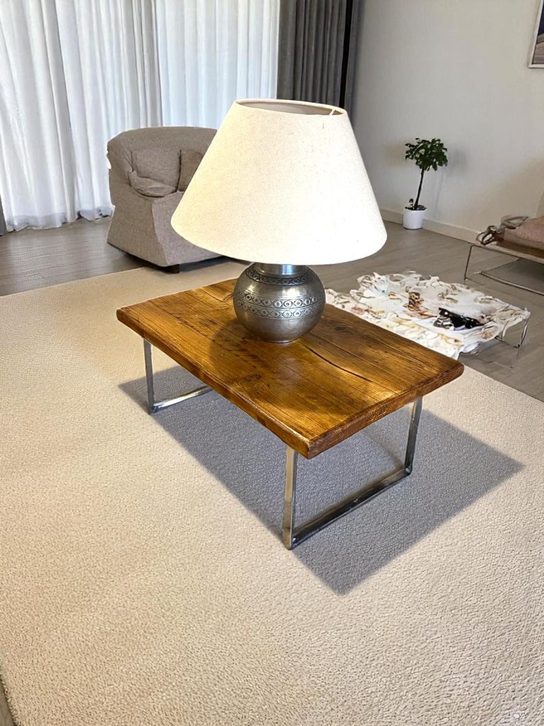 May include: A rectangular wooden coffee table with a natural finish and metal legs. A silver lamp with a cream-colored shade sits on the table. The lamp has a decorative base. The table is on a light-colored rug.