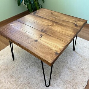 May include: A rectangular wooden coffee table with a dark stain finish. The tabletop is supported by four black hairpin legs. The table is placed on a white, textured rug, with a green wall and plant in the background.