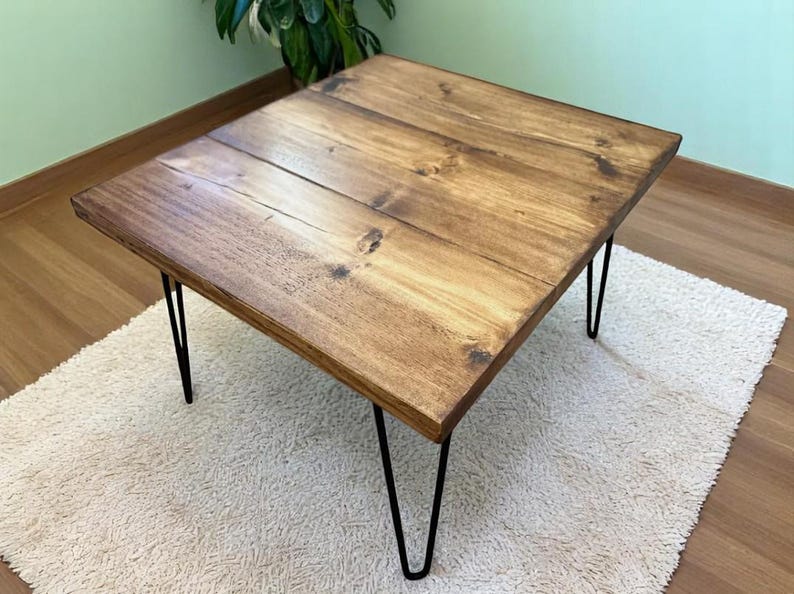 May include: A rectangular wooden coffee table with a dark stain finish. The tabletop is supported by four black hairpin legs. The table is placed on a white, textured rug, with a green wall and plant in the background.
