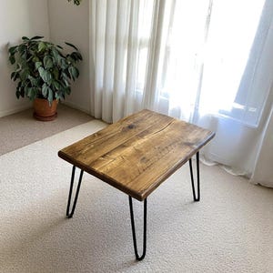 May include: A rectangular wooden coffee table with a natural finish. The table has black hairpin legs. The table is approximately 30 inches long, 18 inches wide, and 16 inches tall.