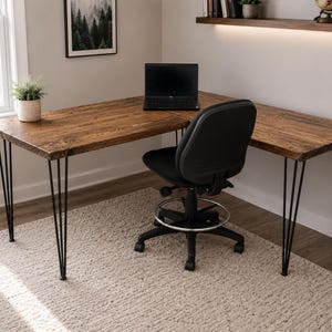 May include: An L-shaped desk with a dark brown wood grain surface and black metal hairpin legs. A black laptop sits on the desk surface. A black office chair with a circular footrest and five-wheel base is positioned in front of the desk.