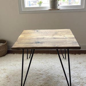 May include: A square wooden table with a natural finish, supported by black hairpin legs. The table is placed on a white, fluffy rug. The wood grain is visible on the tabletop, adding to its rustic charm.