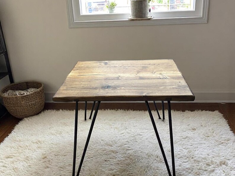 May include: A square wooden table with a natural finish, supported by black hairpin legs. The table is placed on a white, fluffy rug. The wood grain is visible on the tabletop, adding to its rustic charm.